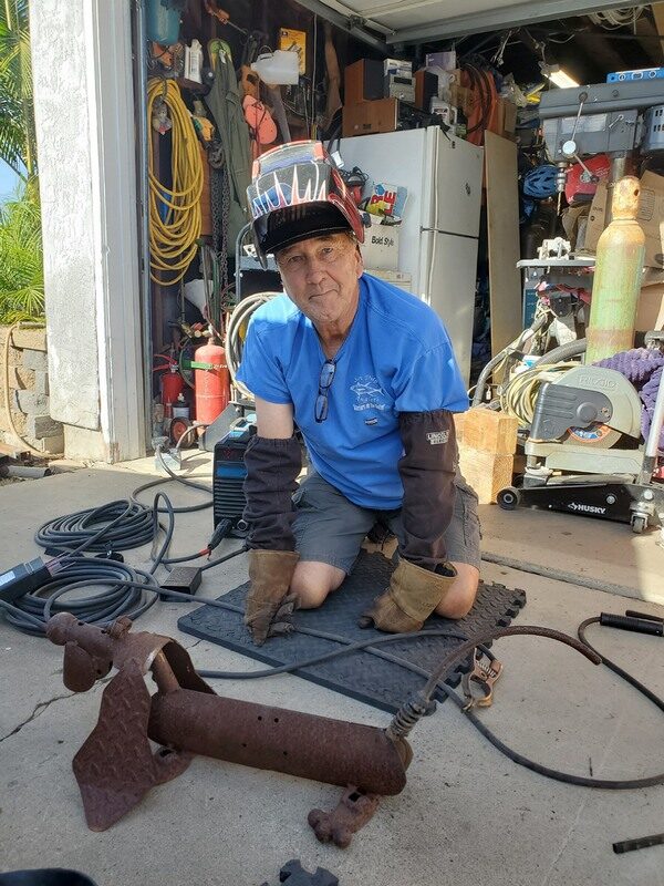 Dave kneeling in his driveway next to a trailer part, welding helmet on